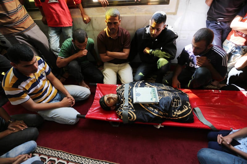 Men sit around the body of a young man wrapped in the flag of the Islamic Jihad faction, lying on a stretcher with a book placed on his chest