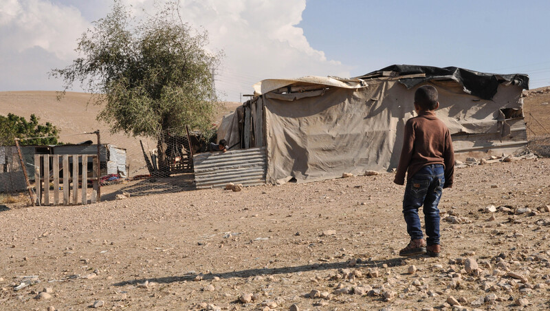 A small child stands in front of a structure made of corrugated sheets and tarps