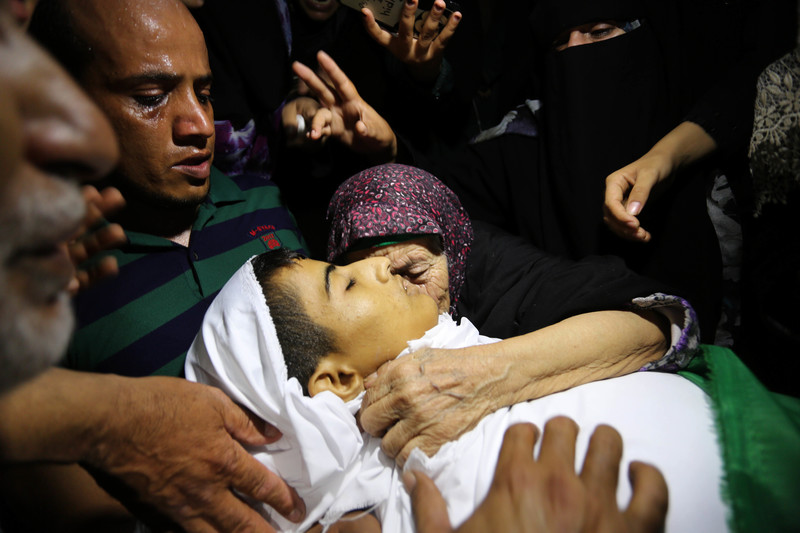 Mourning people surround and place their hands on the shrouded body of slain child