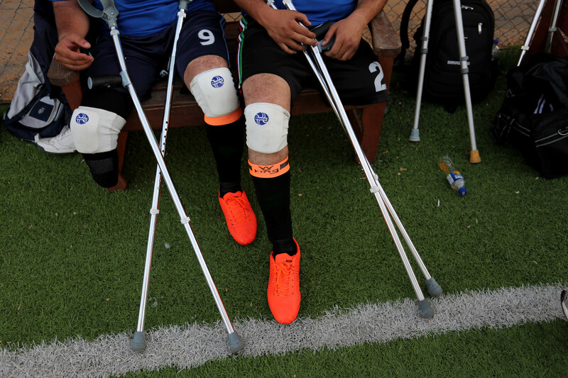 Photo shows legs and crutches of men in football uniform sitting on a bench at a football pitch