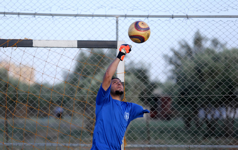 Man wearing football jersey reaches his gloved fist towards a ball approaching the goal net