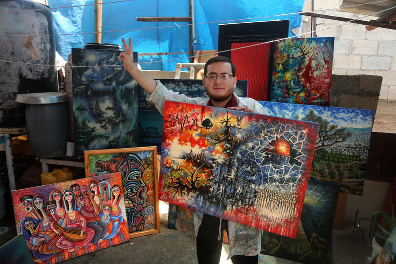 Young man holding large painted canvas gives the victory hand gesture as he stands in a room surrounded by his paintings