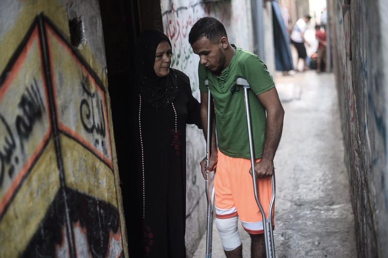 Laila puts her hand on the back of her son Ahmad as he stands using crutches in an alley