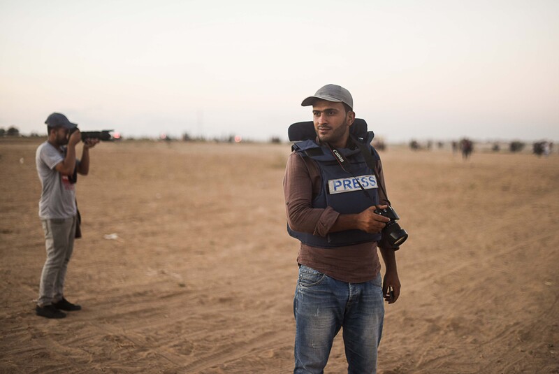 Young man wearing baseball cap and flak jacket holds a camera in front of barren field