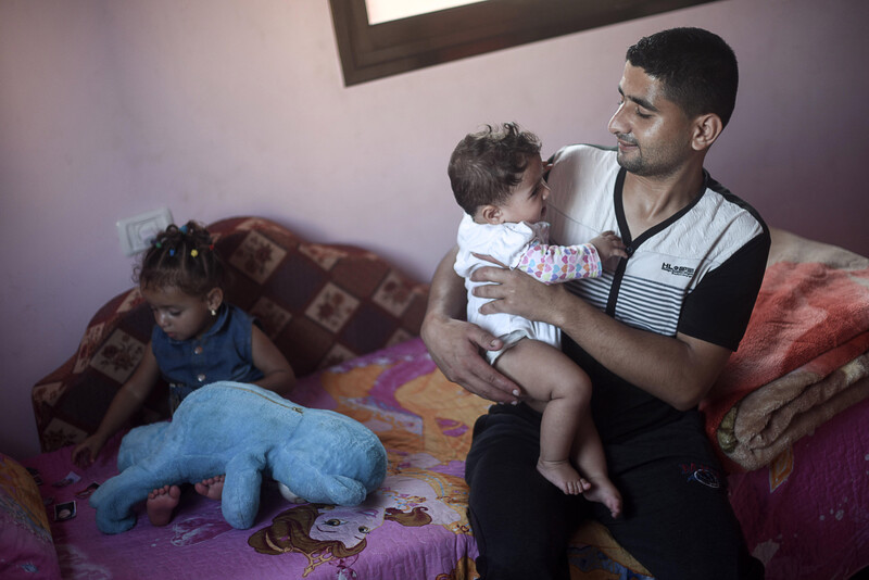 Muhammad al-Najjar smiles and looks at his infant daughter who he is holding while his toddler daughter sits on couch behind him