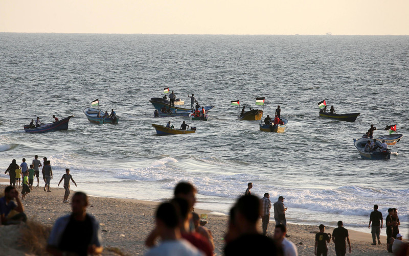 People stand on the beach with boats with Palestinian flags on the water in the background