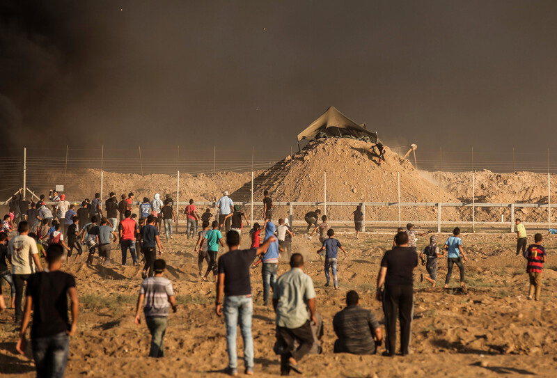 Men and boys stand or run in front of the Gaza boundary fence with an Israeli military installation on a sand hill behind it