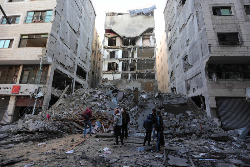 Seven men and boys stand in front of pile of rubble and a damaged multi-story building in densely built-up area