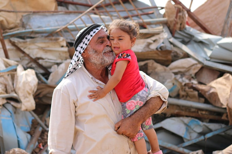 A man with a white beard wearing a kuffiyeh presses his face against a toddler girl while standing in front of rubble