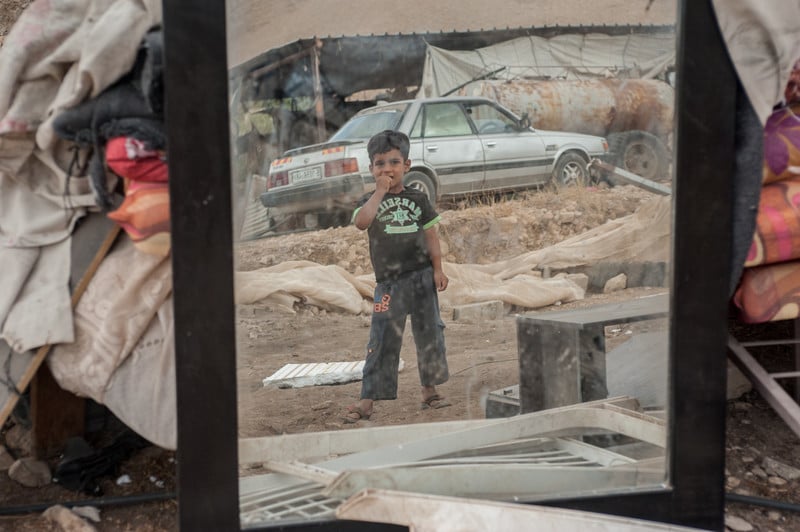 A child is seen in the mirror of a car playing in the rubble of a demolished building.