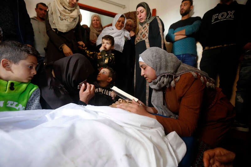 A girl holding a book cradles the face of a shrouded dead man as men, women and children stand nearby