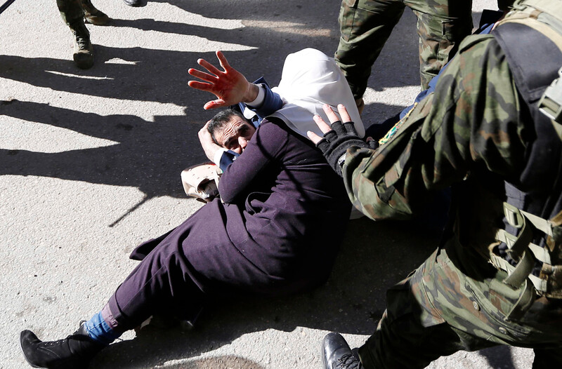 Man lying on ground uses one hand to cover his head and raises his other hand, covered in blood, in the air as a woman lies across him as men in military uniform stand beside them