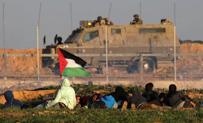 Youths, one holding a Palestine flag, crouch behind a sand embankment with a fence separating them from an Israeli military vehicle and soldiers