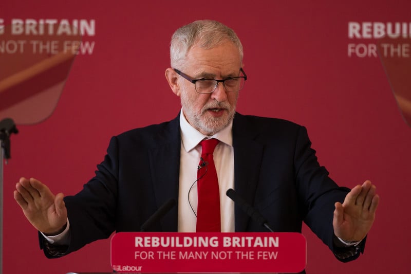 UK opposition leader Jeremy Corbyn standing at a lectern holding his hands up.
