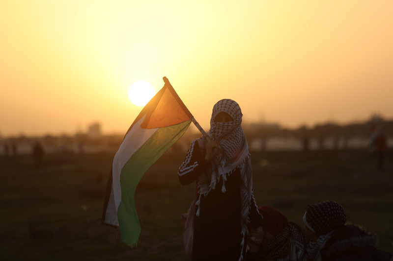 Young woman wearing kuffiyeh around her face holds Palestine flag as sun sets