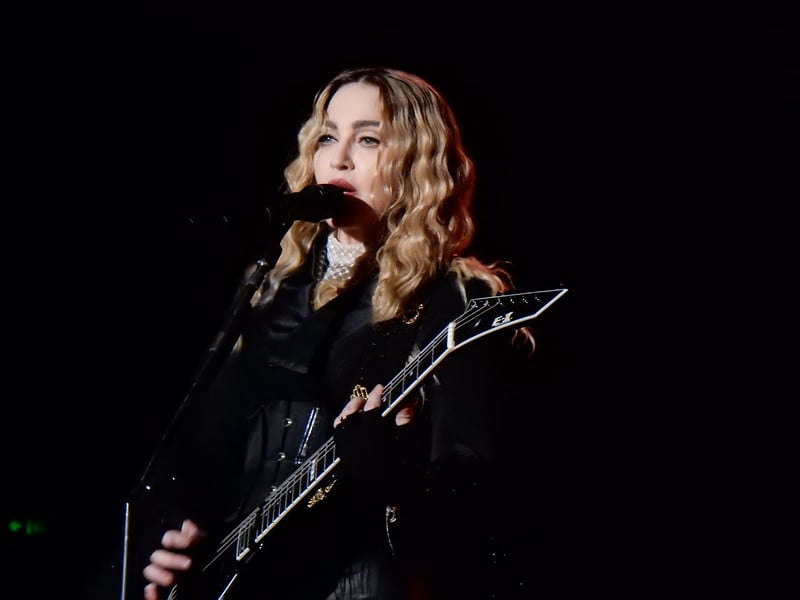 Madonna holding guitar, singing behind a microphone against black background. 