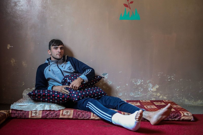 A young man with his right leg in a cast sits on a mattress in front of a bare wall