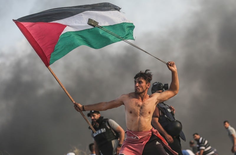 Bare-chested man wields slingshot over his head while holding Palestine flag