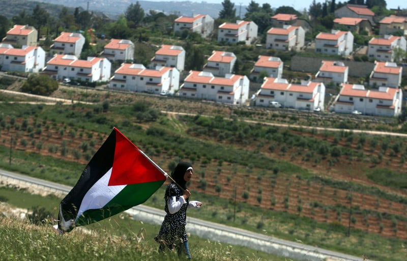 A Palestinian girl holds a Palestinian flag in front of a row of settlement houses.