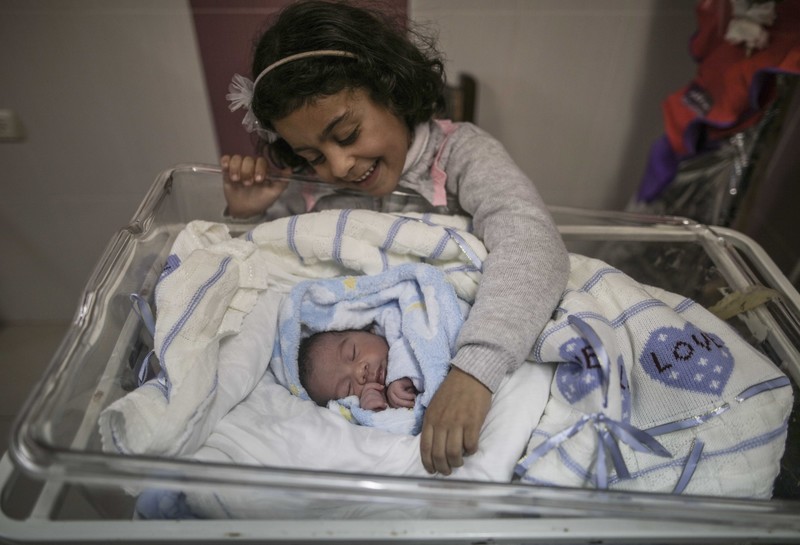 A little girl smiles at a newborn baby wrapped in a blanket.