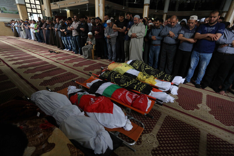 Men stand with their arms folded across their torsos in front of six shrouded bodies