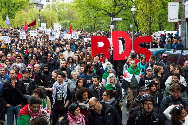 Hundreds of people march, some with signs that say "BDS"