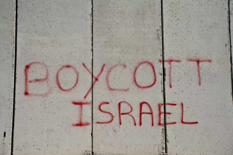 Graffiti written in red on a grey concrete wall reads, "Boycott Israel."