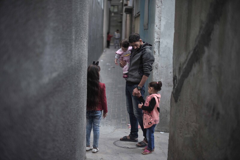 A man carrying a young girl stands flanked by two other children in a narrow grey alley.