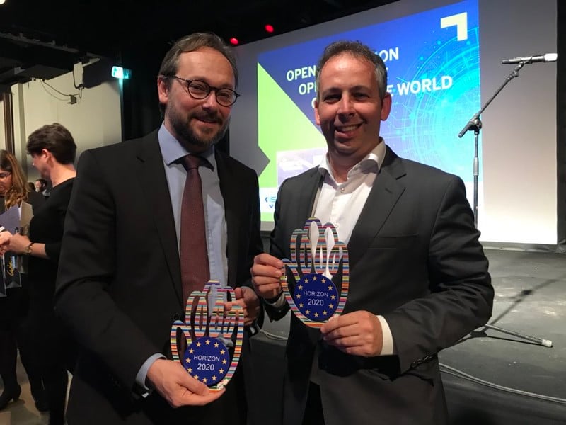 Two men smile and hold trophies that say "Horizon 2020"