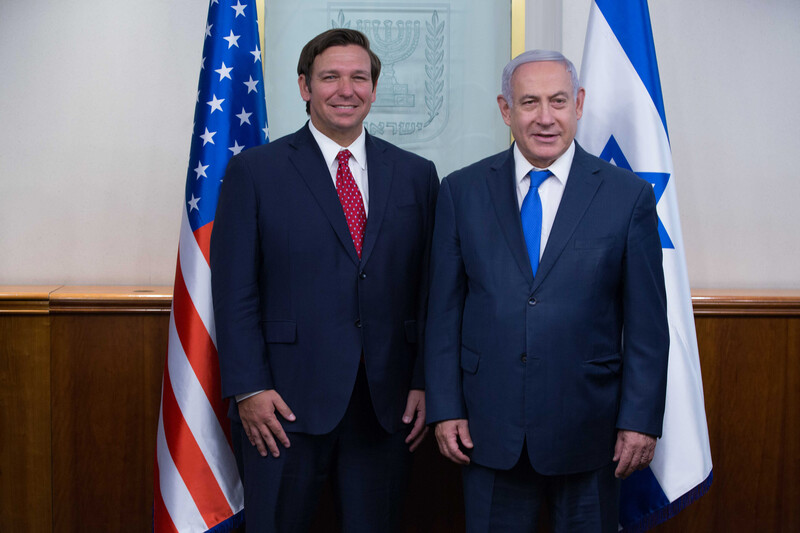 Florida Governor Ron DeSantis stands next to Benjamin Netanyahu, with US and Israel flags behind them.