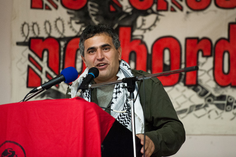 A man wearing a Palestinian scarf speaks at a podium
