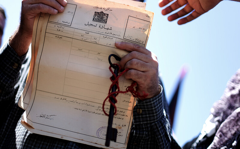 A man's hands with a key and chain wrapped around them holding up a document in Arabic