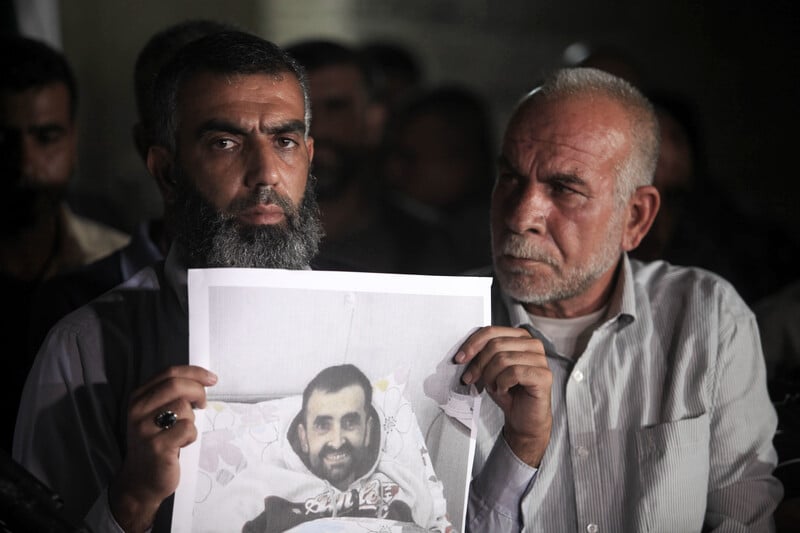  Two men, one of them holds an image of a man in a hospital bed 