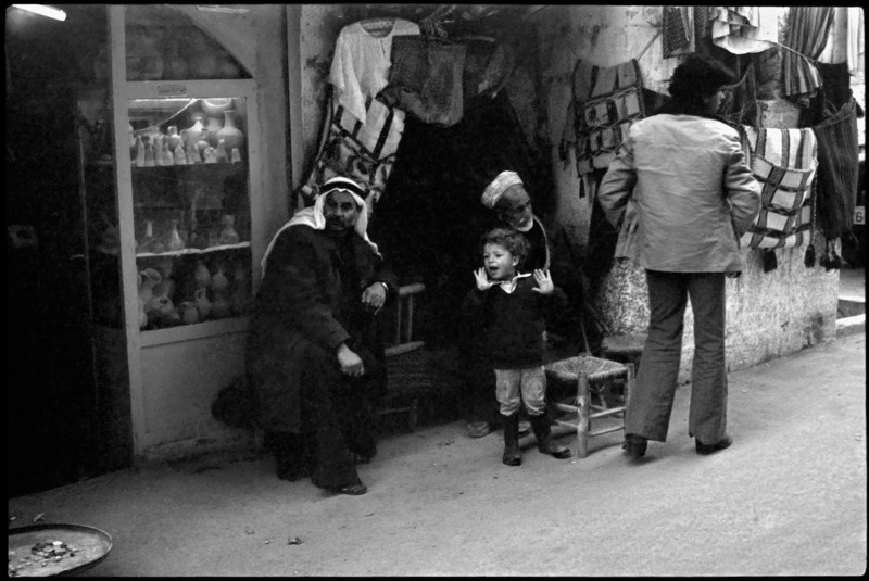 Boy stands next to two men sitting outside souvenir shops