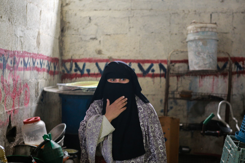 A woman stands in a basic kitchen with her hand held up to her veiled face.