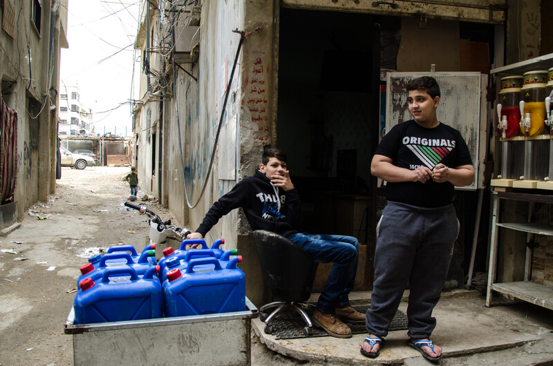 Two boys are next to several large blue plastic water canisters.