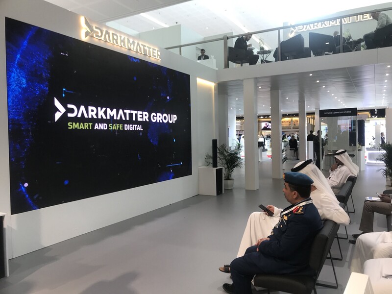 Men sit on chairs facing giant screen