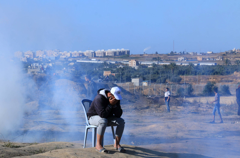 A man sits outside on a plastic chair as smoke billows behind him.