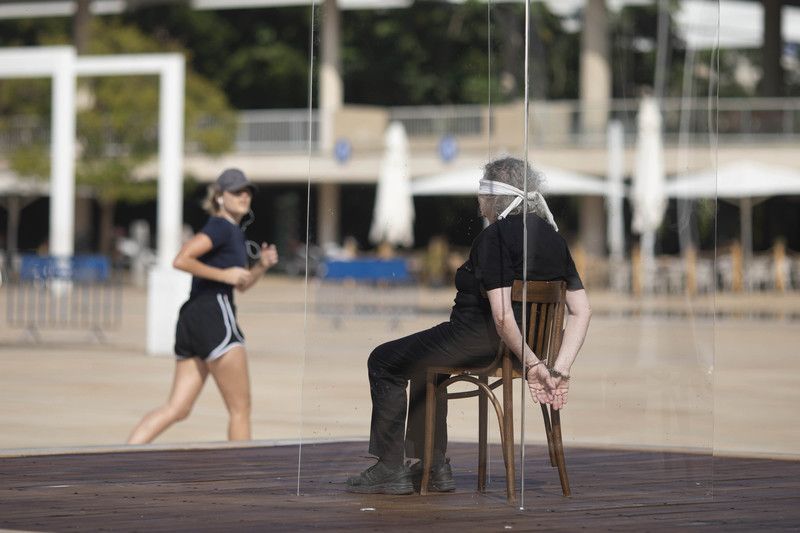 Blindfolded woman cuffed to a chair sits in a glass box 