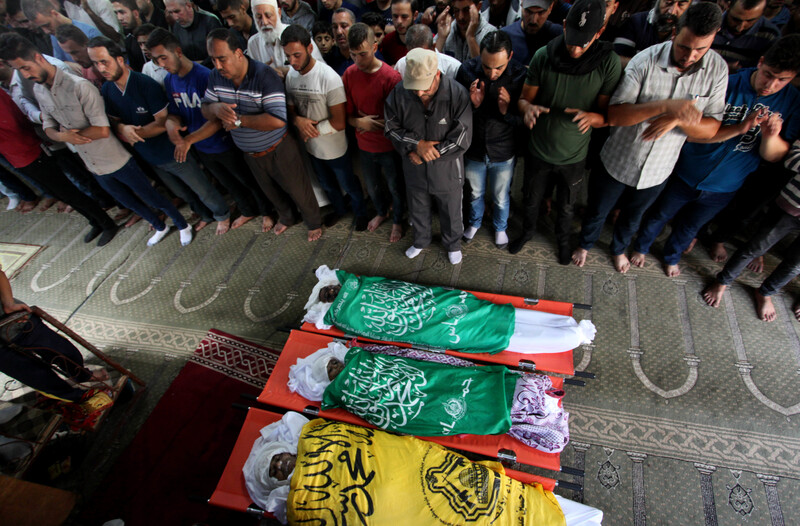 Men stand with their arms folded across their torsos in front of three shrouded bodies