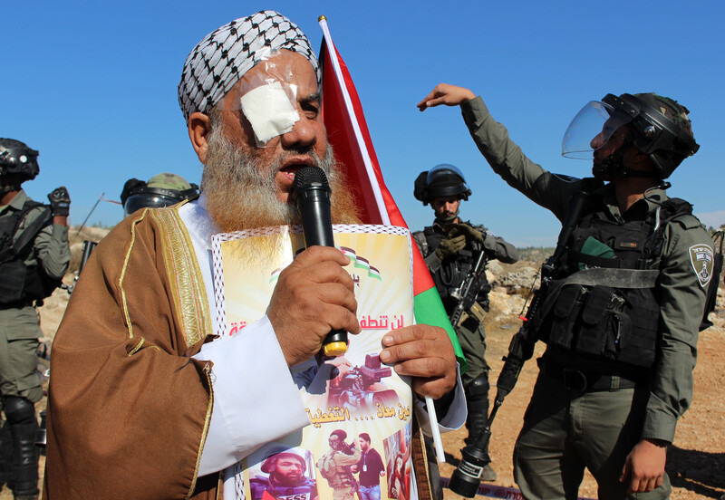 A man with an eyepatch speaks into a microphone while a soldier gesticulates behind him