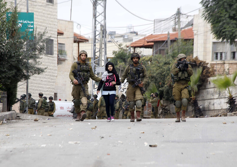 Two armed soldiers walk with a young girl between them while a third points a rifle somewhere in the distance