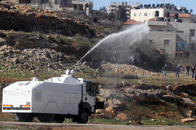 A white armored vehicle sprays liquid at some people on a hillside in the distance