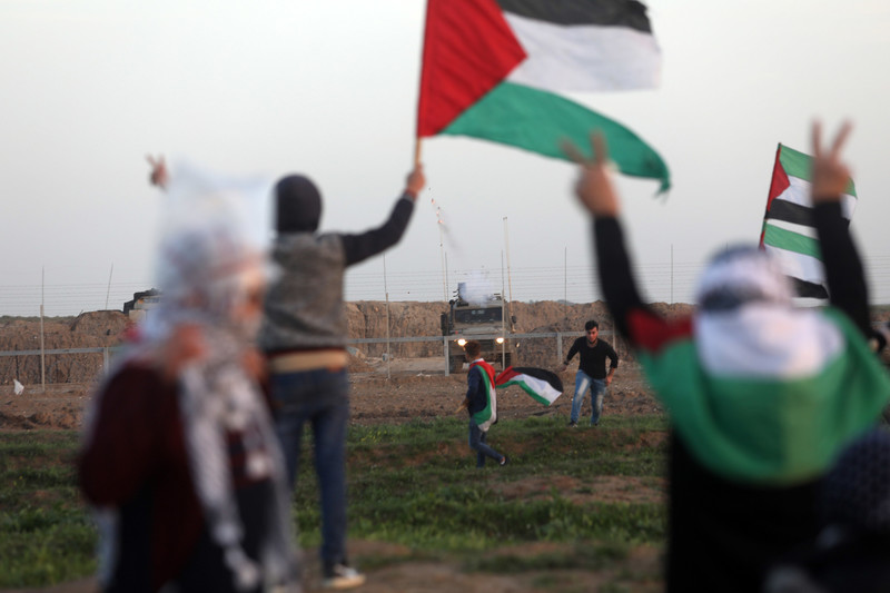 Palestinians hold flags at boundary fence with Israeli army jeep on opposite side
