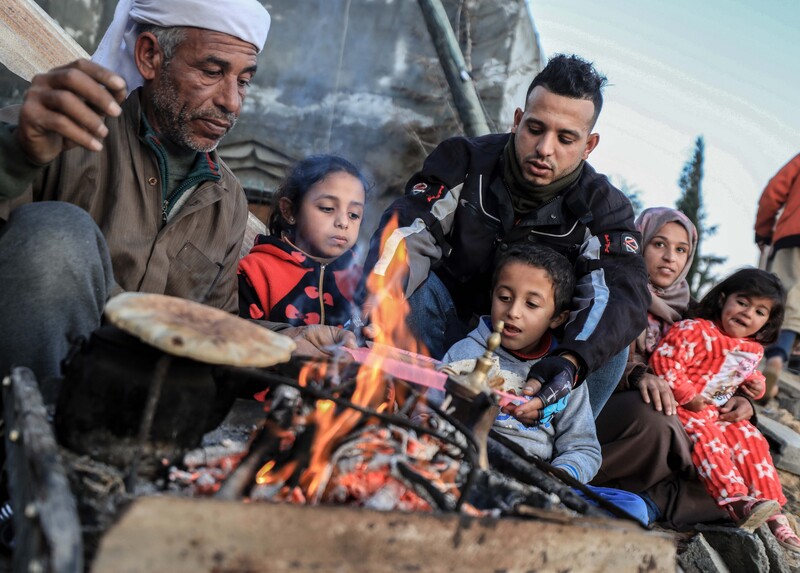 Three adults and three children warm bread around an open fire.