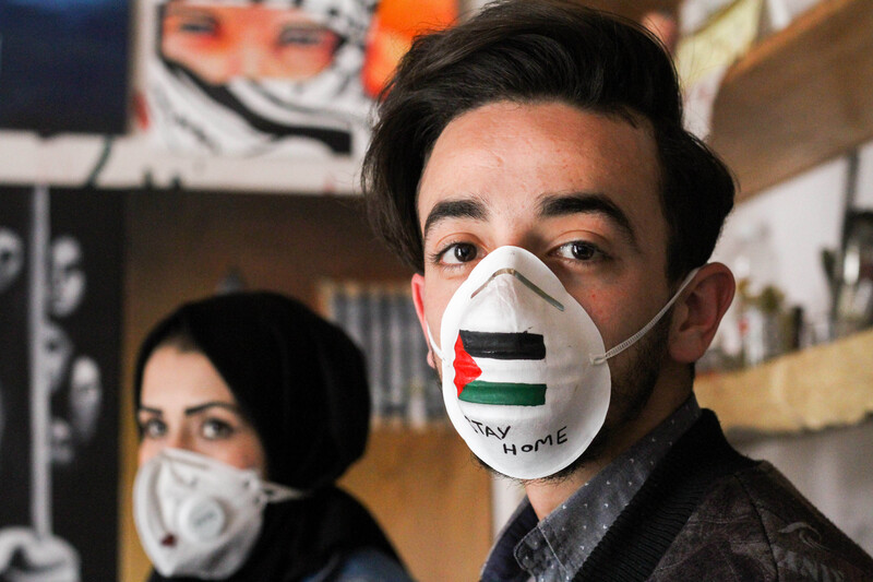 A man and a woman wear painted face masks, one bearing the Palestinian flag and the words: "Stay at home".