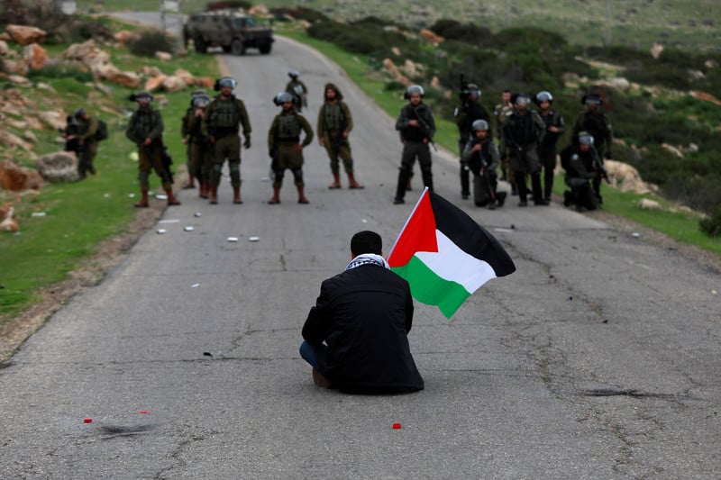 A man waving a flag sits in the middle of a road facing a line of armed soldiers