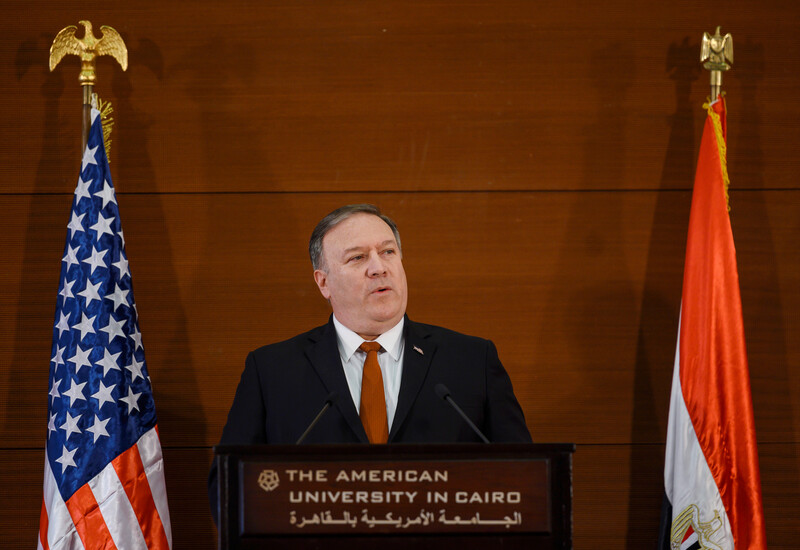 Man in suit stands before podium between two flags 