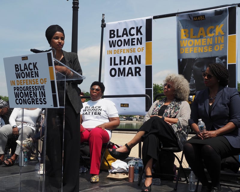 Woman speaks at podium with women and banners behind her