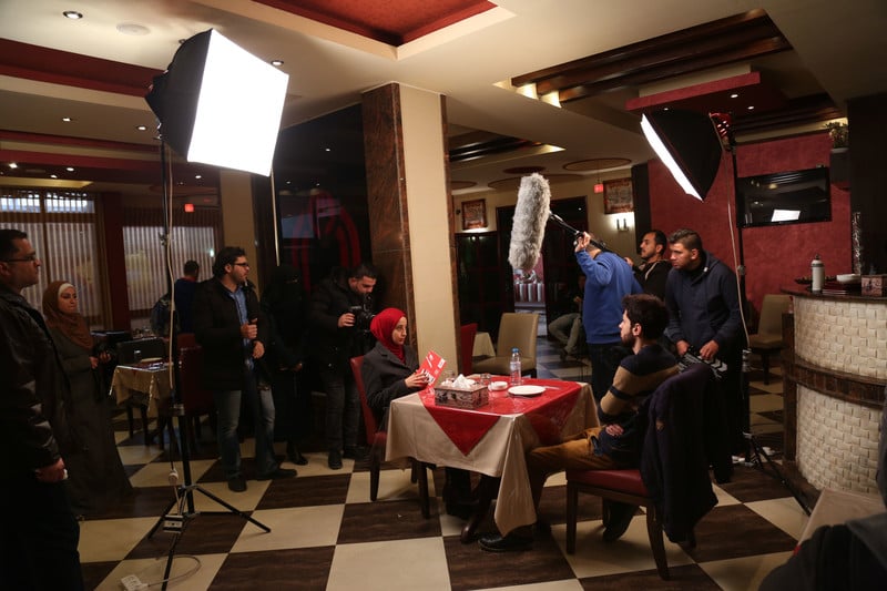Film crew stands around male and female actors seated across from each other at restaurant table